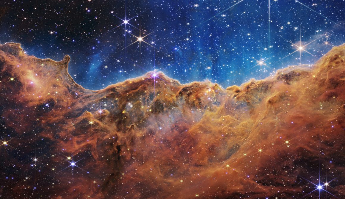 What looks much like craggy mountains on a moonlit evening is actually the edge of a nearby, young, star-forming region NGC 3324 in the Carina Nebula. Captured in infrared light by the Near-Infrared Camera (NIRCam) on NASA’s James Webb Space Telescope, this image reveals previously obscured areas of star birth. Called the Cosmic Cliffs, the region is actually the edge of a gigantic, gaseous cavity within NGC 3324, roughly 7,600 light-years away. The cavernous area has been carved from the nebula by the intense ultraviolet radiation and stellar winds from extremely massive, hot, young stars located in the center of the bubble, above the area shown in this image. The high-energy radiation from these stars is sculpting the nebula’s wall by slowly eroding it away. NIRCam – with its crisp resolution and unparalleled sensitivity – unveils hundreds of previously hidden stars, and even numerous background galaxies. Several prominent features in this image are described below. • The “steam” that appears to rise from the celestial “mountains” is actually hot, ionized gas and hot dust streaming away from the nebula due to intense, ultraviolet radiation. • Dramatic pillars rise above the glowing wall of gas, resisting the blistering ultraviolet radiation from the young stars. • Bubbles and cavities are being blown by the intense radiation and stellar winds of newborn stars. • Protostellar jets and outflows, which appear in gold, shoot from dust-enshrouded, nascent stars. • A “blow-out” erupts at the top-center of the ridge, spewing gas and dust into the interstellar medium. • An unusual “arch” appears, looking like a bent-over cylinder. This period of very early star formation is difficult to capture because, for an individual star, it lasts only about 50,000 to 100,000 years – but Webb’s extreme sensitivity and exquisite spatial resolution have chronicled this rare event. Located roughly 7,600 light-years away, NGC 3324 was first cat