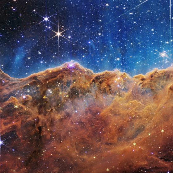 What looks much like craggy mountains on a moonlit evening is actually the edge of a nearby, young, star-forming region NGC 3324 in the Carina Nebula. Captured in infrared light by the Near-Infrared Camera (NIRCam) on NASA’s James Webb Space Telescope, this image reveals previously obscured areas of star birth. Called the Cosmic Cliffs, the region is actually the edge of a gigantic, gaseous cavity within NGC 3324, roughly 7,600 light-years away. The cavernous area has been carved from the nebula by the intense ultraviolet radiation and stellar winds from extremely massive, hot, young stars located in the center of the bubble, above the area shown in this image. The high-energy radiation from these stars is sculpting the nebula’s wall by slowly eroding it away. NIRCam – with its crisp resolution and unparalleled sensitivity – unveils hundreds of previously hidden stars, and even numerous background galaxies. Several prominent features in this image are described below. • The “steam” that appears to rise from the celestial “mountains” is actually hot, ionized gas and hot dust streaming away from the nebula due to intense, ultraviolet radiation. • Dramatic pillars rise above the glowing wall of gas, resisting the blistering ultraviolet radiation from the young stars. • Bubbles and cavities are being blown by the intense radiation and stellar winds of newborn stars. • Protostellar jets and outflows, which appear in gold, shoot from dust-enshrouded, nascent stars. • A “blow-out” erupts at the top-center of the ridge, spewing gas and dust into the interstellar medium. • An unusual “arch” appears, looking like a bent-over cylinder. This period of very early star formation is difficult to capture because, for an individual star, it lasts only about 50,000 to 100,000 years – but Webb’s extreme sensitivity and exquisite spatial resolution have chronicled this rare event. Located roughly 7,600 light-years away, NGC 3324 was first cat